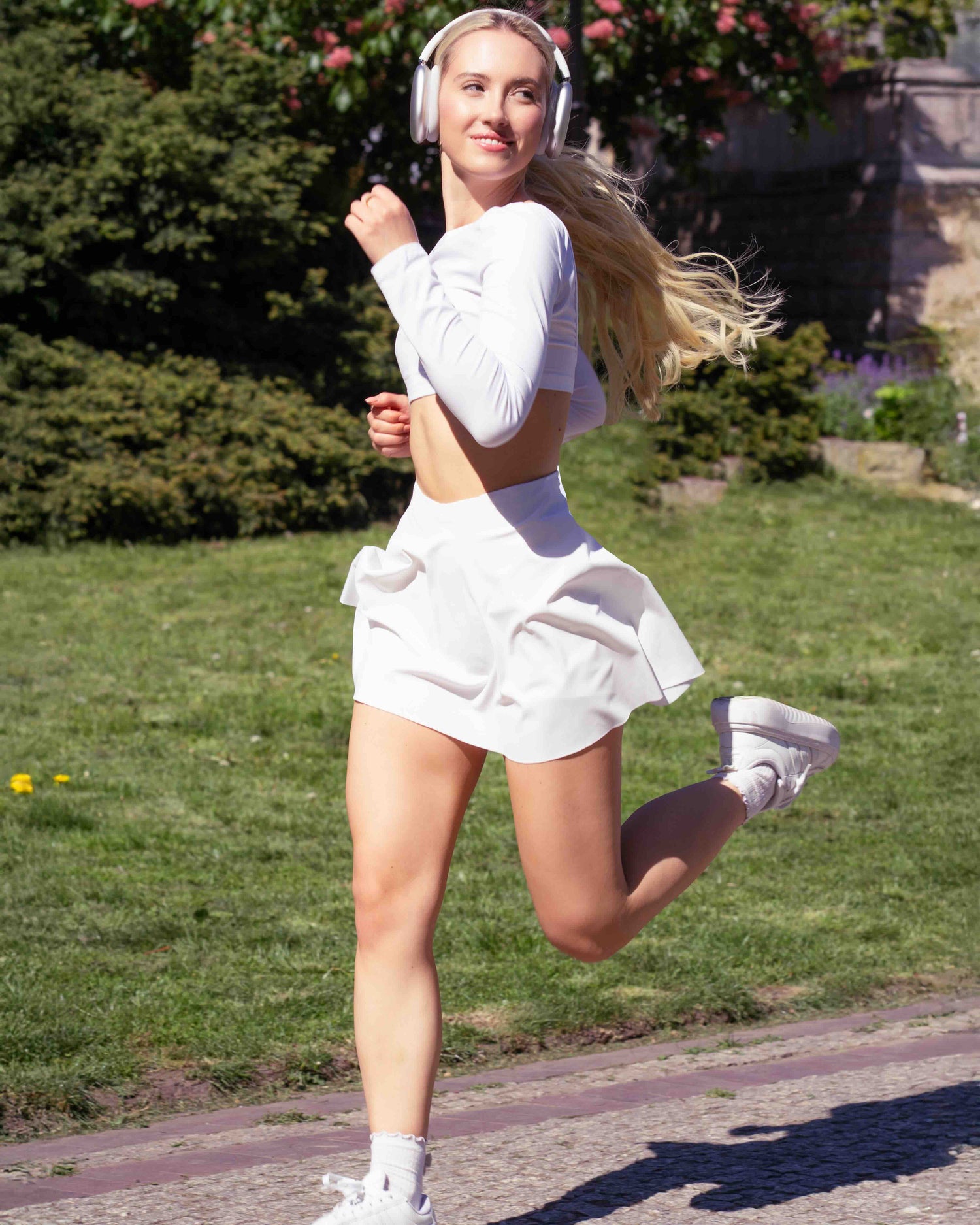 Uśmiechnięta kobieta w białej sportowej spódniczce Glissmove z wbudowanymi spodenkami biegnie w parku. Ubrana w biały, długi crop top i słuchawki bezprzewodowe. Połączenie kobiecego stylu i sportowej funkcjonalności.
